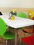 Wnętrze dziennego domu. Jadalnia. Biały stół, na blacie którego stoi kolorowa ozdoba w kształcie kwiatów. Kolorowe krzesła wokół stołu.